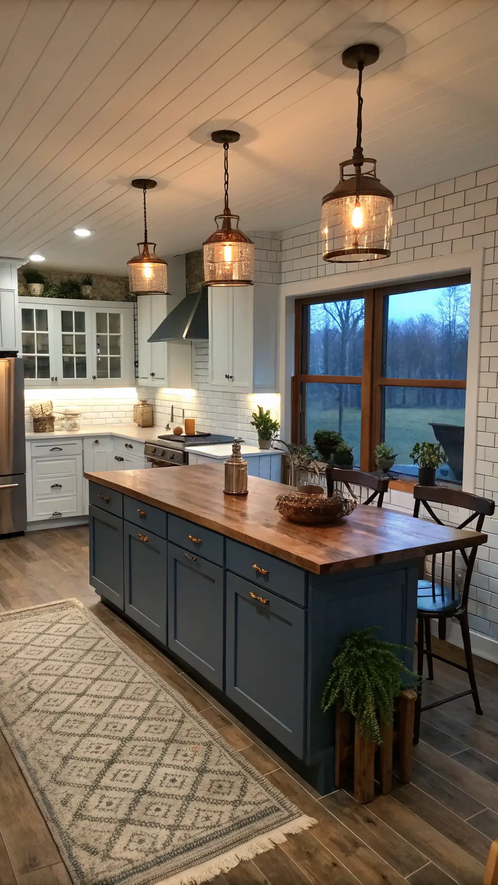 Twilight view of a modern farmhouse kitchen with slate blue aluminum cabinets, reclaimed wood island top, vintage copper pendants, and a white subway tile backdrop, captured from the dining area showcasing the entire space and cozy mixed lighting.