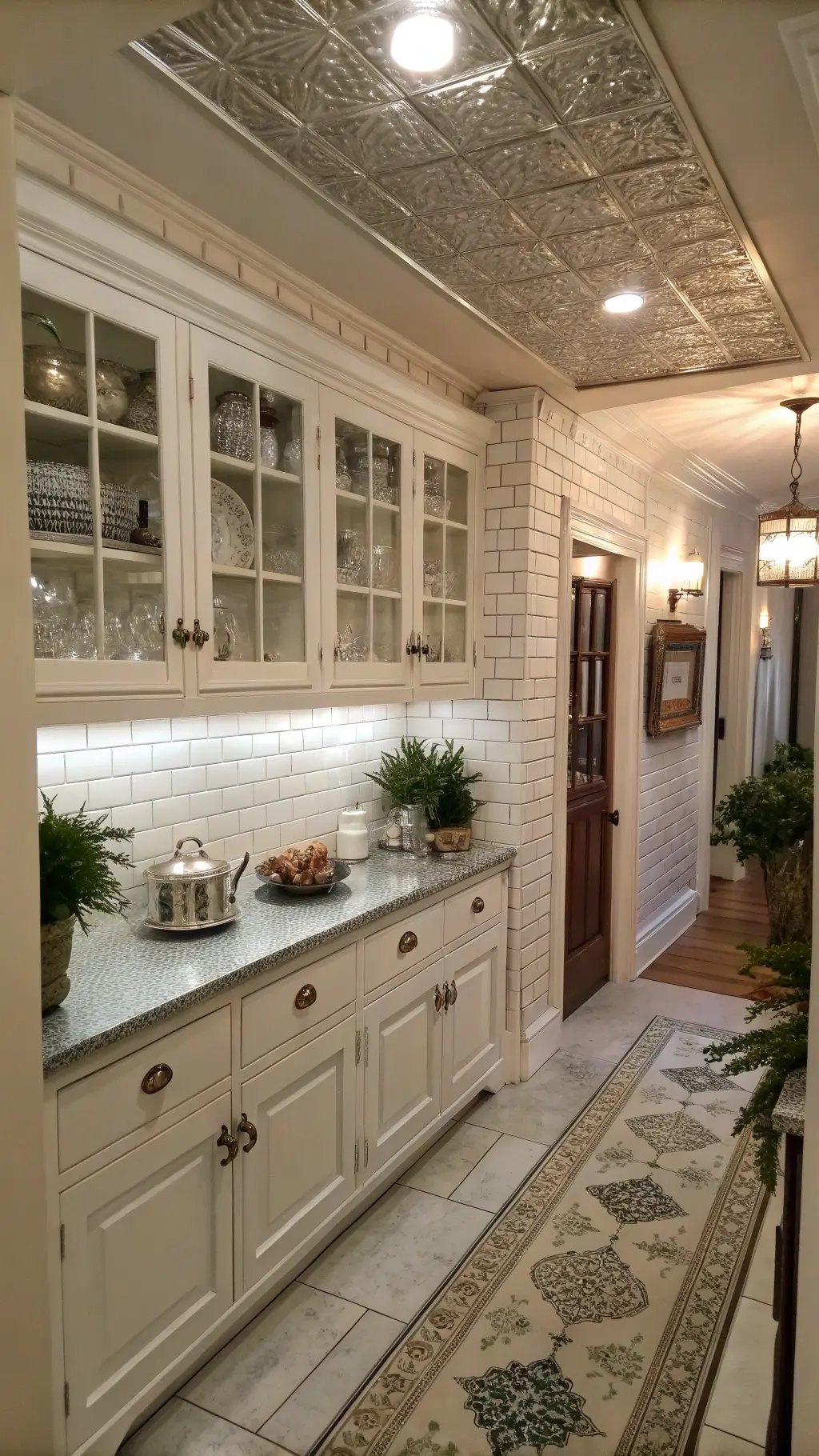 Antique white kitchen with pressed tin ceiling, marble tiles, crystal knobs on cabinets, silver tea service and topiaries in zinc planters shot through a doorway.