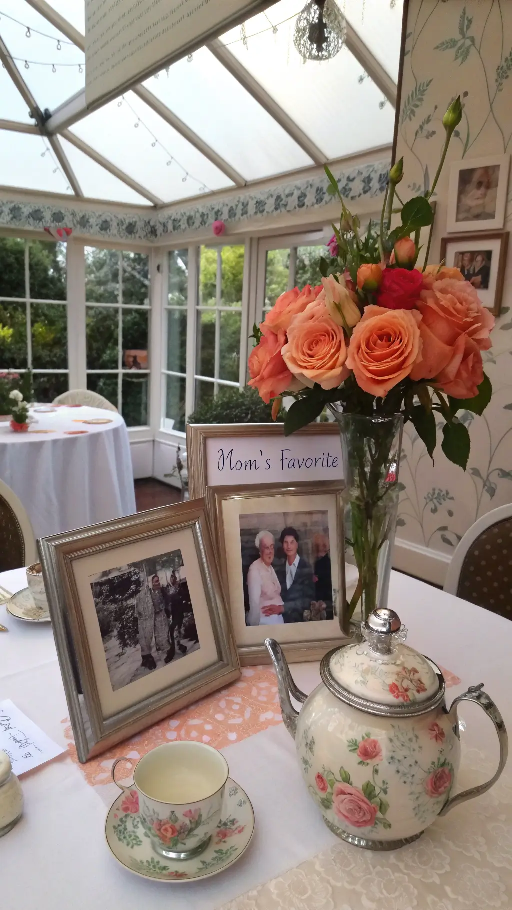 Aerial view of a sunlit conservatory dining area adorned with family photos in silver frames, garden roses in a vintage teapot vase, and handwritten notes tucked between flowers, set against a botanical wallpaper backdrop.
