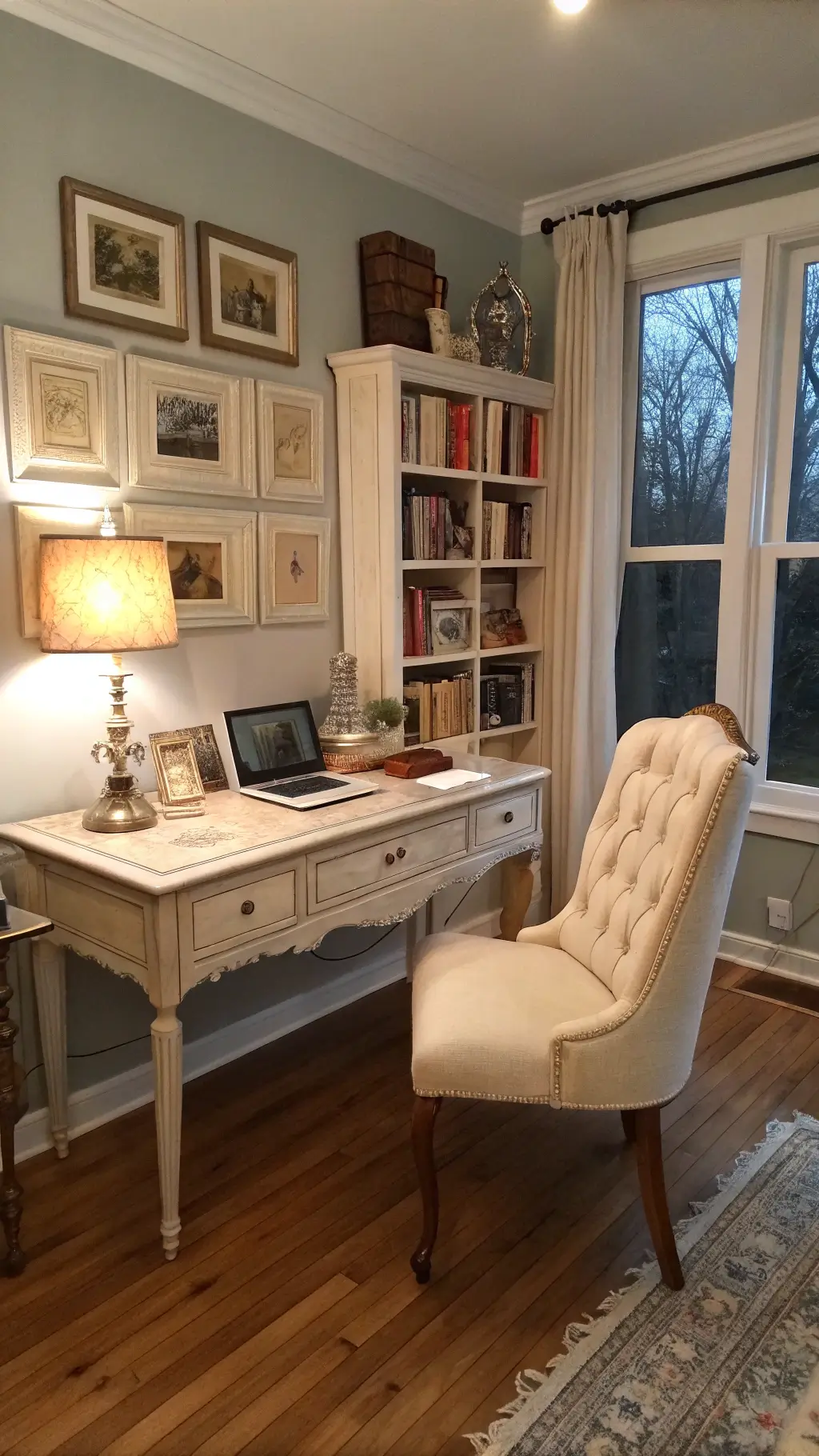 Shabby chic home office with French provincial desk, tufted linen chair, built-in bookshelf filled with vintage books, crystal lamp, and gilded frames collection on pale gray walls.