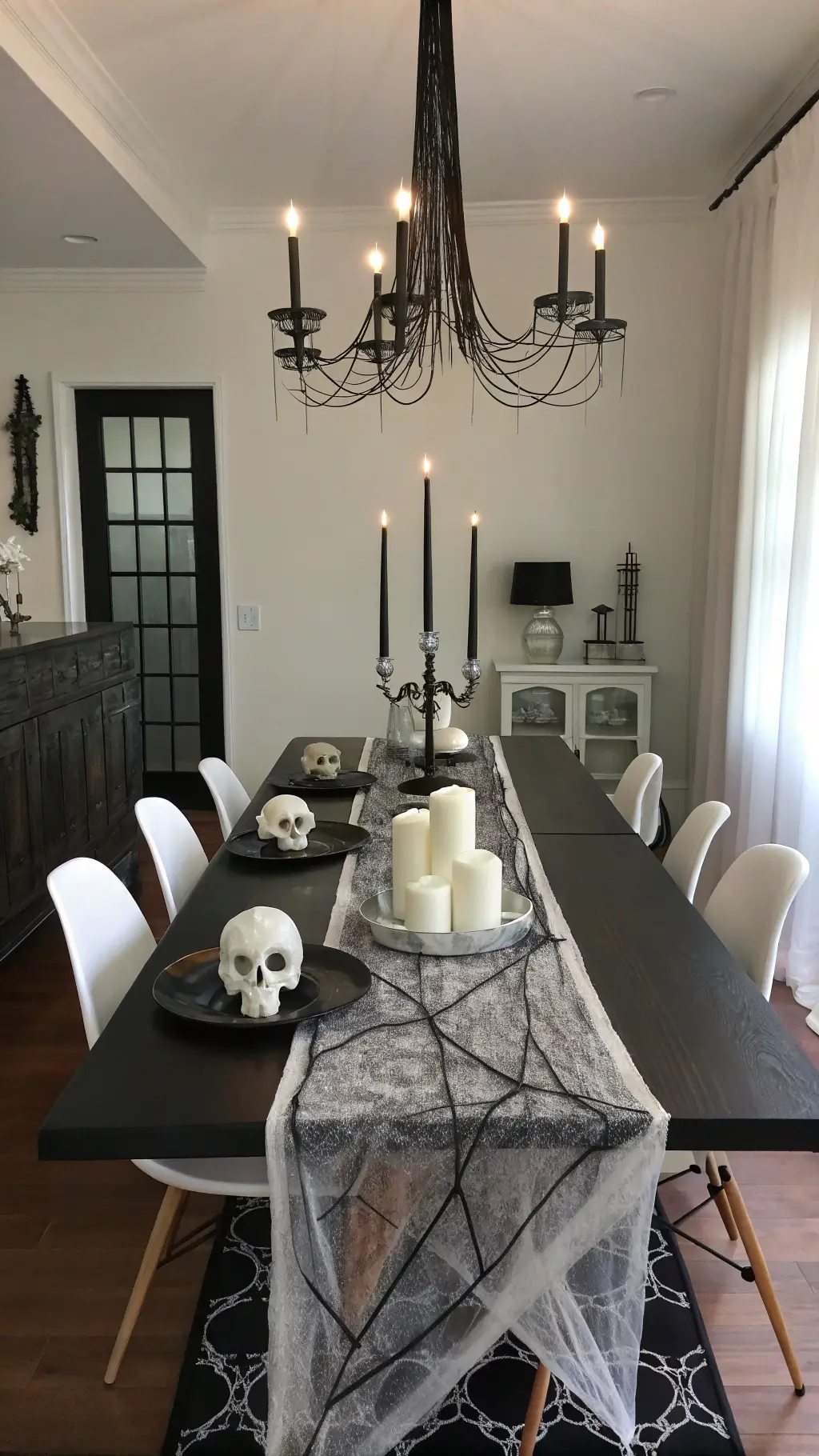 Overhead view of a modern minimalist dining room at midnight featuring a black table with dark gauze runner and silver spider webs, lit by tall black taper candles and a metal chandelier draped with cobwebs, which is surrounded by ghost chairs and white ceramic skull centerpieces.