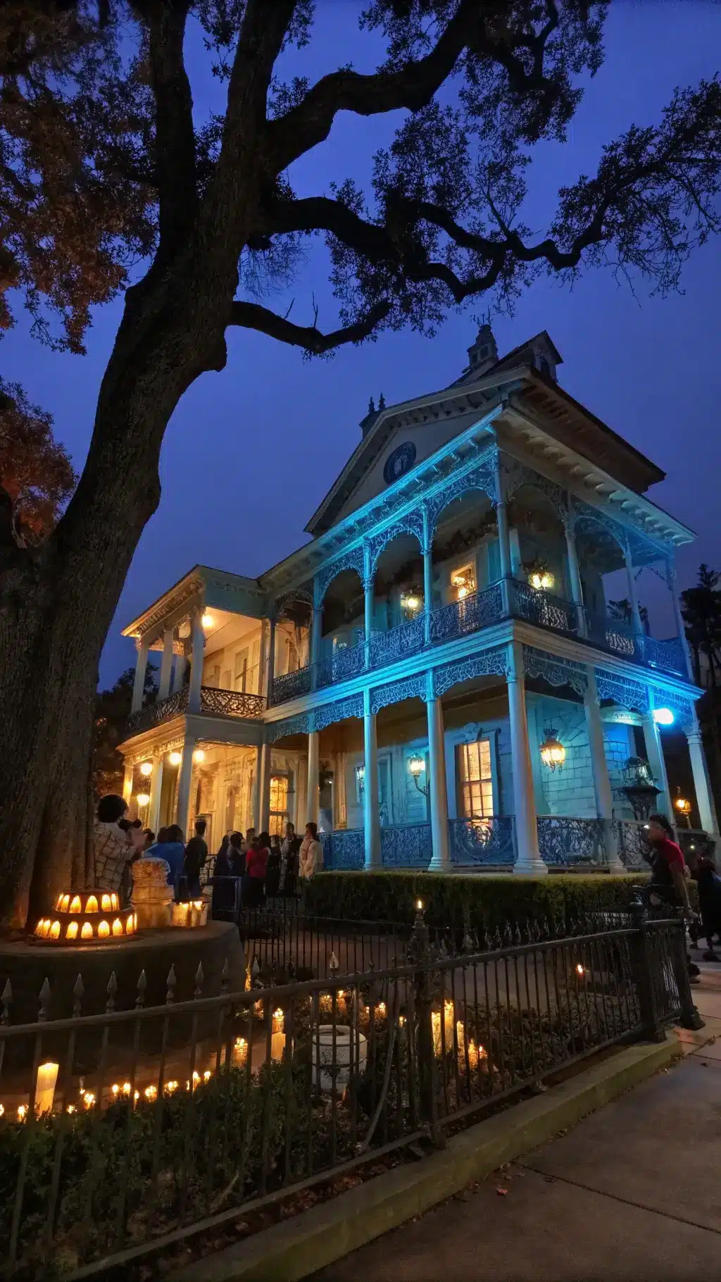 Haunted mansion facade illuminated under blue moonlight, featuring ghostly figures projected in windows, a deceased oak tree adorned with floating candles, and an animatronic raven perched on black wrought iron fence, captured with a tilt-shift lens for a miniature effect.