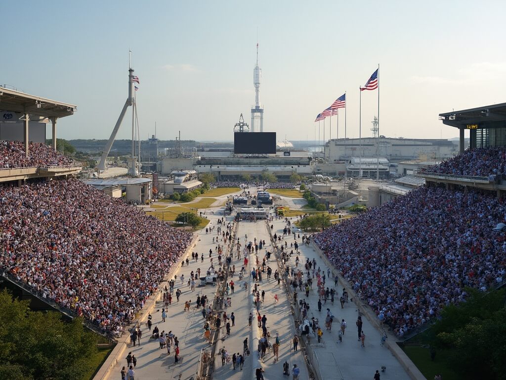 Aerial view of crowded Kennedy Space Center Visitor Complex during a major launch event with spectators in bleachers, Vehicle Assembly Building, rocket garden, interactive exhibits, tour buses, and American flags with anticipation for the Artemis mission launch.